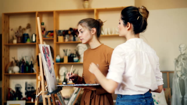 Beautiful-young-woman-artist-is-teaching-her-student-to-paint-flowers-working-in-studio-together-with-oil-paints-and-palette.-Visual-arts-and-youth-concept.