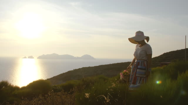 Beautiful-young-woman-wearing-fashion-colorful-dress-with-skirt-and-hat-carrying-flowers-in-basket-at-sunset-on-Ponza-Island-mountain-Italy.