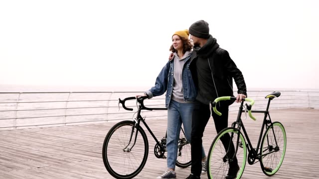 Moderne-junge-Hipster-Paare,-die-zusammen-mit-ihren-Fahrrädern-am-Meer-am-Herbsttag-zu-umarmen.-Fuß-durch-Holzdeck-tagsüber,-Lächeln-und-reden.-Slow-motion