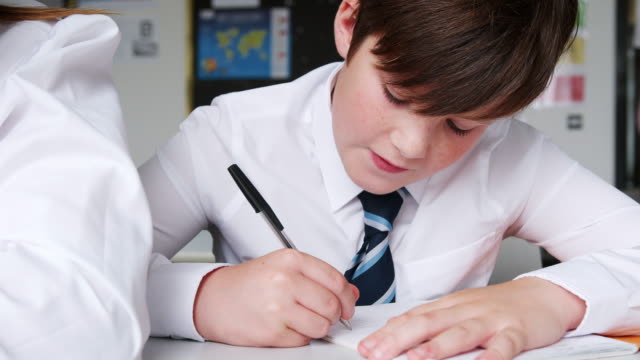 Estudiante-de-secundaria-de-hombre-usando-escritura-uniforme-en-ejercicio-libro-sentado-en-el-escritorio-en-el-aula
