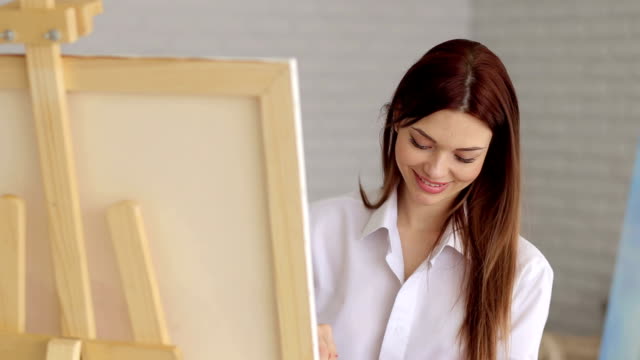 Ein-junges-sexy-Mädchen-in-einem-weißen-Hemd-stützt-sich-auf-Leinwand-im-Atelier-für-Zeichnung.-Kunst