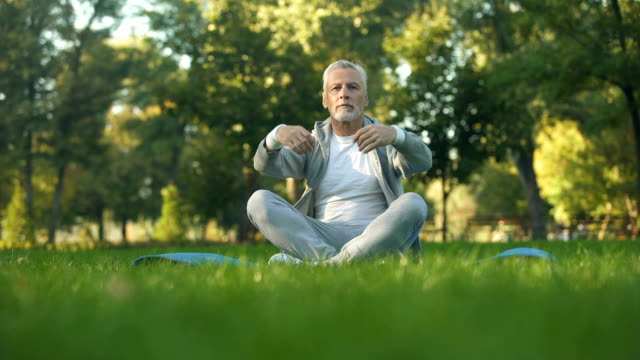 Healthy-senior-man-meditating-in-park,-sitting-lotus-pose-on-yoga-mat,-zen