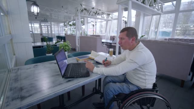 Online-Bildung,-erfolgreiche-studentische-ungültig-Männer-auf-Rollstuhl-verwendet-moderne-Notebooktechnologie-lernen-aus-Büchern-Notizen-im-Notizbuch-close-up-sitzen-am-Tisch-und-Online-Lehre