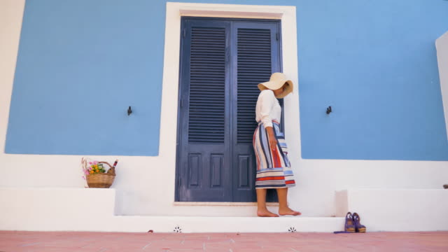 Young-happy-woman-with-bicycle-walking-in-front-of-blue-house-door-patio.-Fashion-white-shirt,-large-hat,-colorful-skirt-and-sunglasses.-Ponza-island,-italy.