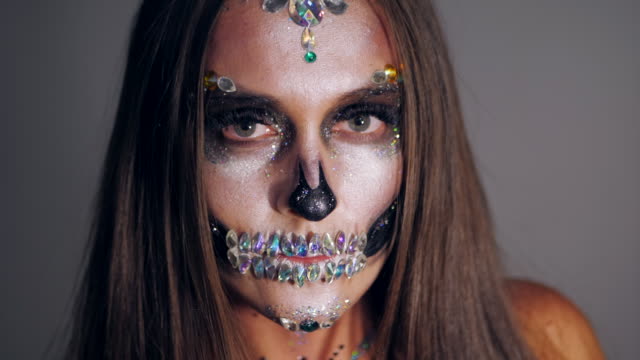 Young-girl-with-creative-halloween-face-art-in-the-studio-with-grey-background.-Portrait-of-glamorous-skull-with-rhinestones-and-sequins.-Professional-make-up-for-the-celebration.-Slow-motion