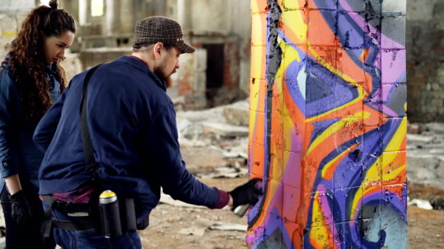 Urban-artist-handsome-bearded-man-is-teaching-amateur-student-to-work-with-aerosol-paint-while-decorating-old-industrial-building-with-abstract-graffiti.