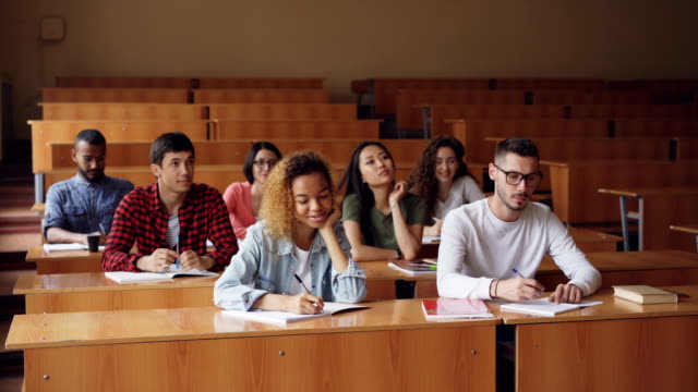 Group-of-students-is-listening-to-teacher-and-writing-sitting-at-tables-in-classroom-while-professor-is-talking-and-gesturing.-Higher-education-and-people-concept.