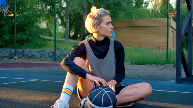 Beautiful-blonde-female-basketball-player-sitting-on-ground-and-playing-with-ball,-park-in-background