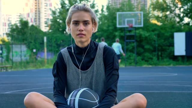 Smiling-young-female-basketball-player-listening-music-through-earphones-and-sitting-on-court,-men-playing-on-background-in-park