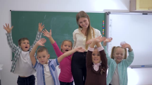 Escuela-primaria,-grupo-de-divertidos-niños-saltando-y-agitando-las-manos-cerca-de-la-maestra-en-el-fondo-de-la-Junta