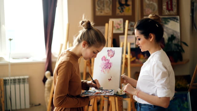 Pretty-woman-painting-teacher-is-teaching-young-girl-to-mix-paints-on-palette-creating-beautiful-color-during-art-class-in-nice-modern-studio.-People-and-education-concept.
