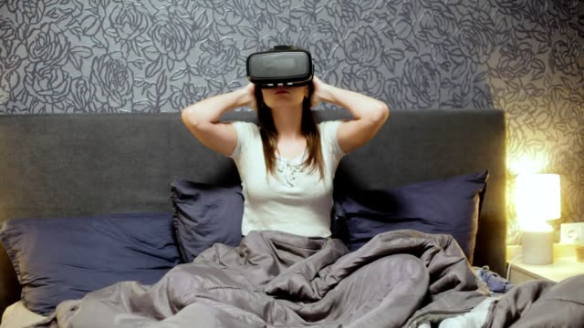 Schöne-junge-Frau-trägt-VR-Kopfhörer-auf-dem-Bett.-Umzusehen,-Lächeln-und-mit-Gesten-mit-den-Händen.