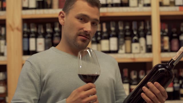 Portrait-of-handsome-male-who-smelling-wine-glass.-A-guy-with-a-bottle-of-wine-and-a-wine-glass-in-his-hands-standing-on-the-background-of-shelves-with-alcoholic-beverages.
