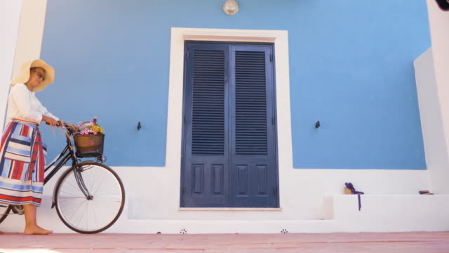 Mujer-joven-feliz-con-bicicleta-caminando-frente-a-patio-de-la-puerta-de-la-casa-azul.-Camisa-de-moda-blanco,-gran-sombrero,-falda-colorida-y-gafas-de-sol.-Isla-de-Ponza,-Italia.