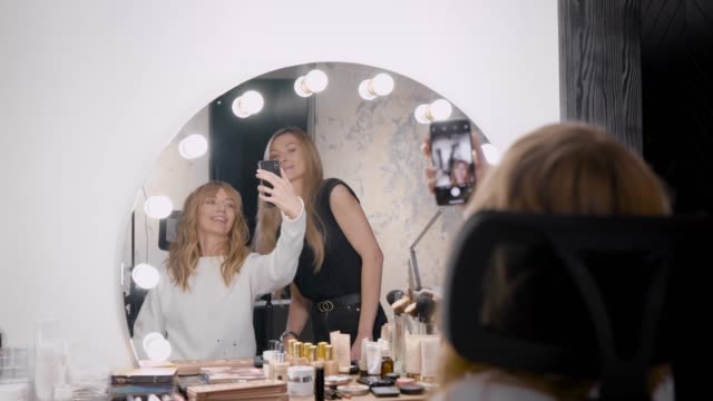 female-client-of-beauty-shop-is-taking-selfie-with-professional-makeup-artist-in-front-of-mirror-with-lamps