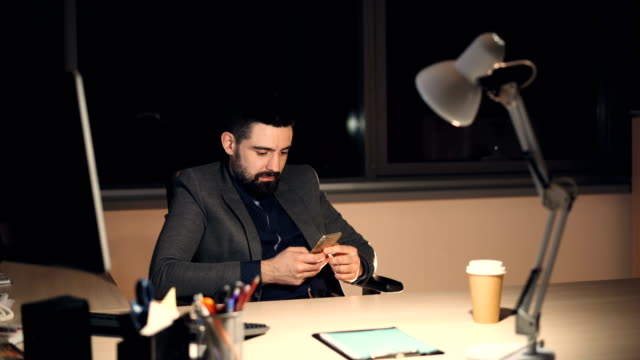 Gutaussehender-bärtiger-junger-Mann-nutzt-Smartphone-Bildschirm-berühren-und-lächelnd,-spät-in-der-Nacht-allein-im-Büro-arbeiten.-Kommunikation-und-moderne-Technologie-Konzept.