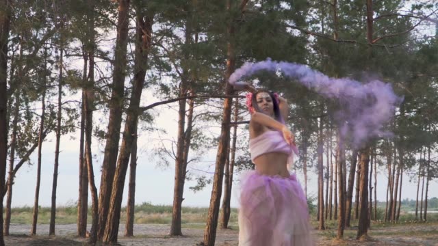 Charmante-Frau-mit-hellen-Make-up-in-einem-rosa-Kleid-tanzen-mit-Rauchbomben-auf-dem-Hintergrund-der-Pinien.-Der-Tanz-von-einem-sinnlichen-Mädchen-mit-einer-wunderschönen-Frisur-mit-Blumen.-Slow-Motion.
