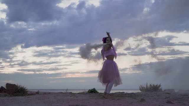Tender-young-woman-with-brilliance-makeup-in-a-pink-dress-dancing-with-smoke-bombs-outdoors.-The-dance-of-a-sensual-girl-with-a-wonderful-hairstyle-with-flowers.-Slow-motion.