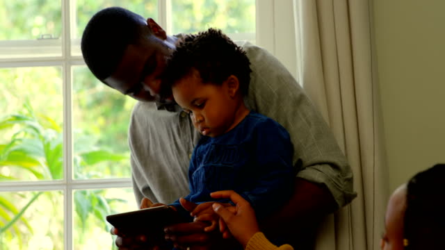 Vista-frontal-de-padre-e-hijo-negro-usando-digital-y-sentado-en-el-alféizar-de-la-ventana-de-la-casa-confortable-4k