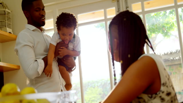 Vista-frontal-del-joven-padre-negro-sosteniendo-a-su-hijo-y-hablando-con-la-madre-en-la-cocina-en-casa-4k