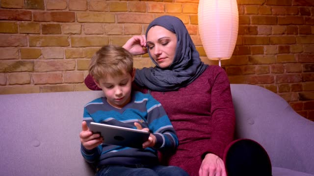 Aufmerksamer-kleiner-Junge-spielt-Spiel-auf-Tablet-und-seine-muslimische-Mutter-in-Hijab-beobachten-seine-Tätigkeit-zu-Hause.