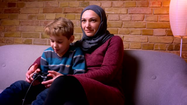 Konzentrierter-kleiner-Junge-und-seine-muslimische-Mutter-im-Hijab-spielen-Videospiel-mit-Joystick-zusammen-zu-Hause.