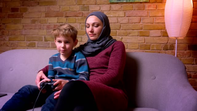 Konzentrierter-kleiner-Junge,-der-Videospiel-spielt,-und-seine-muslimische-Mutter-im-Hijab-versucht,-Joystick-zu-holen,-um-zu-versuchen,-zu-spielen.