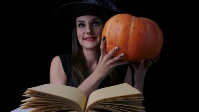 Attraktive-Hexe-auf-schwarzem-Hintergrund-vor-einem-offenen-Zauberbuch,-blickt-hinter-einem-Kürbis-hervor-und-lächelt-jedes-Mal,-wenn-sich-ihre-Stimmung-ändert,-Halloween-Konzept.