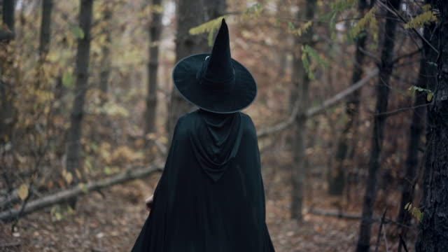 Unrecognizable-woman-as-black-witch-walks-between-trees-in-autumn-forest.Girl-in-long-dress,-cape,-fairy-hat.-Halloween-concept,-cosplay-dressing-up