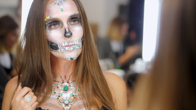 Young-girl-with-creative-halloween-face-art-looking-into-mirror-reflection-in-the-dressing-room.-Portrait-of-glamorous-skull-with-rhinestones-and-sequins.-Professional-make-up-for-the-celebration
