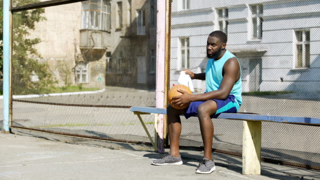 Sad-Afro-American-male-sitting-on-the-bench-and-playing-ball,-loneliness