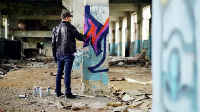 Bearded-young-man-is-painting-graffiti-inside-old-industrial-buliding-using-aerosol-paint,-he-is-making-bright-abstract-image-on-column.-Creativity-and-people-concept.
