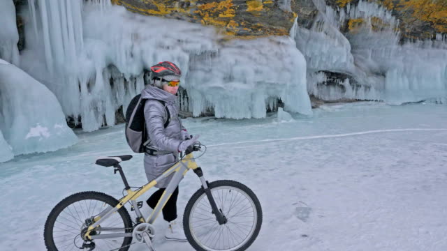 Woman-is-walking-beside-bicycle-near-the-ice-grotto.-The-rock-with-ice-caves-and-icicles-is-very-beautiful.-The-girl-is-dressed-in-silvery-down-jacket,-backpack-and-helmet.-The-traveler-is-ride-cycle.