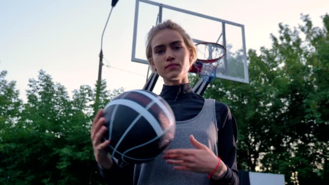 Jugador-de-baloncesto-femenina-rubia-atractiva-jugando-con-la-pelota-y-mirando-a-cámara,-en-el-parque,-durante-el-día