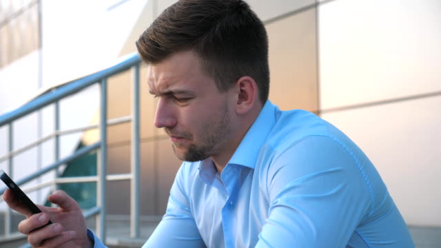 Profile-of-desperate-young-businessman-talking-on-smart-phone-and-sitting-on-stairs-near-office-building.-Close-up-of-upset-sad-manager-got-a-very-bad-news-on-cell-phone-and-crying-outdoor.-Side-view