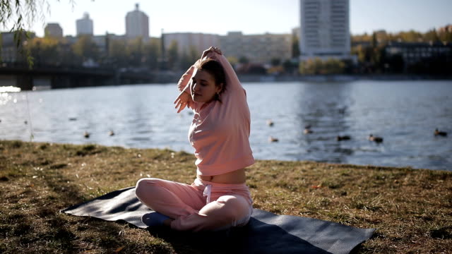 Mädchen-in-einen-Trainingsanzug-an-den-Ufern-eines-städtischen-Flusses-macht-Yoga-auf-einer-speziellen-Matte-und-hört-Musik.