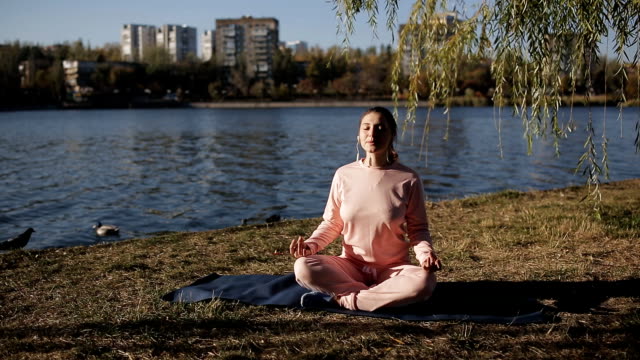 Girl-in-a-tracksuit-on-the-banks-of-an-urban-river-does-yoga-on-a-special-Mat-and-listens-to-music.