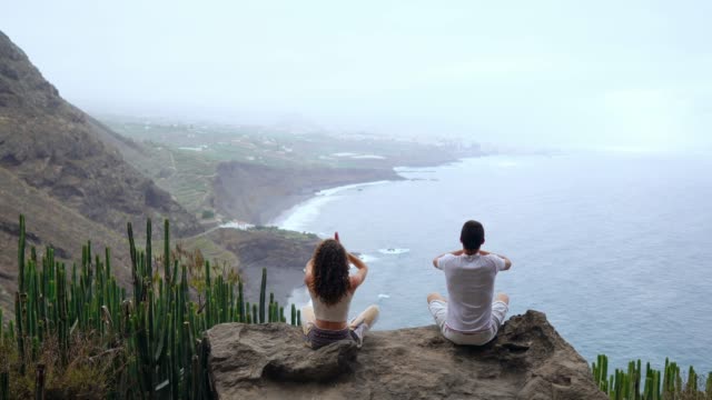 Ein-Mann-und-eine-Frau-sitzen-auf-einem-Berg,-der-Blick-auf-den-Ozean-sitzt-auf-einem-Stein-hob-ihre-Hände-und-die-Durchführung-eines-entspannenden-Atems-zu-meditieren.-Kanarische-Inseln