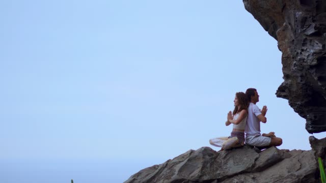 Man-and-woman-sitting-on-top-of-a-mountain-on-a-rock-back-to-back-meditate-and-do-yoga-on-the-background-of-the-ocean.
