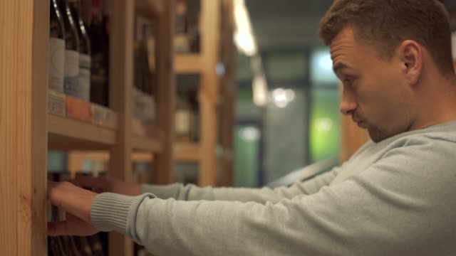 Portrait-of-cheerful-man-is-taking-white-wine-bottle-in-the-shop-close-up-Customer-is-choosing-drink-in-alcohol-shop
