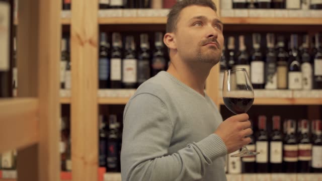 Handsome-man-standing-with-glass-of-red-wine-in-liquor-store.-Woman-in-the-background-is-choosing-a-wine-bottle