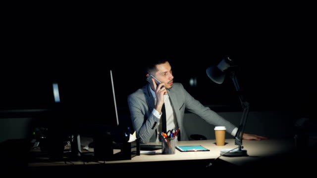 Jungen-Mann-in-spät-in-die-Nacht-telefonieren-mit-Handy-dann-lachend-sitzen-am-Schreibtisch-im-Büro-arbeiten,-betonte.-Einzelnen-Computer,-Kaffee-zum-Mitnehmen-sind-sichtbar.