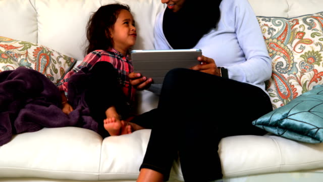 Young-mother-using-a-digital-tablet-with-her-daughter-on-the-sofa-at-home-4k