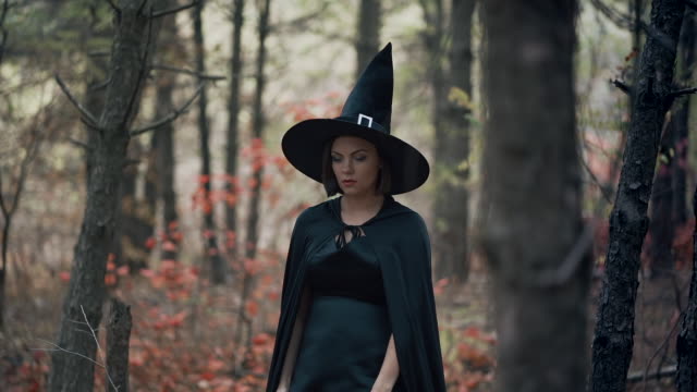 Woman-as-black-witch-walks-between-trees-in-autumn-forest.Girl-in-long-dress,-cape,-fairy-hat.-Halloween-concept,-cosplay-dressing-up