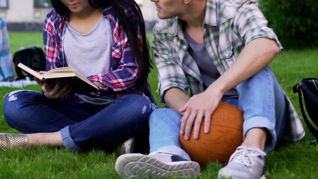 Students-sitting-on-lawn-together,-girl-reading-and-guy-talking-to-her,-flirt
