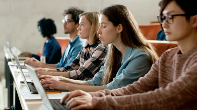 Fila-de-Multi-étnica-estudiantes-trabajando-en-las-computadoras-portátiles-mientras-se-escucha-a-una-conferencia-en-el-aula-moderna.-Luminoso-estudio-en-la-Universidad-de-los-jóvenes.