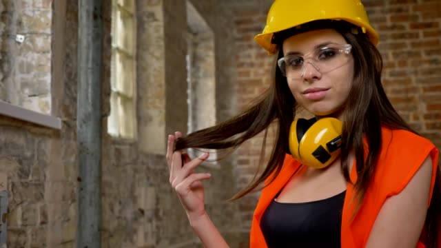 Young-pretty-attractive-women-in-helmet,-earphones-and-glasses-looking,-pointing-in-camera,-touching-her-long-hair,-smiling,-female-builder,-brick-building-background