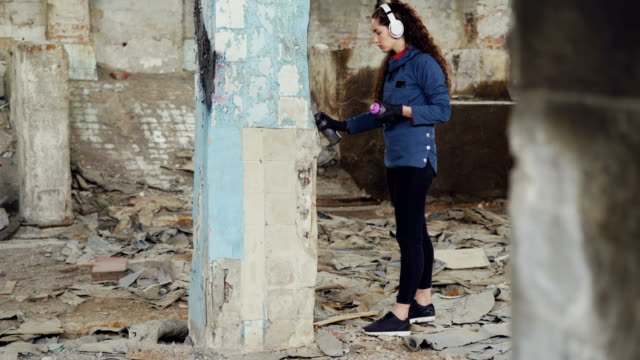 Tilt-up-shot-of-slim-girl-modern-painter-drawing-graffiti-on-high-column-in-ruined-warehouse-with-aerosol-paint-and-listening-to-music-with-headphones.