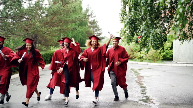 Aufgeregte-Absolventen-mit-einem-Diplom-auf-dem-Campus-Territorium-tragen-Kleider-und-Trachtenhüte-laufen,-es-regnet.-Höhere-Bildung-und-Glück-Konzept.