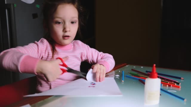Cute-little-preschool-European-girl-child-in-pink-sweater-cutting-her-drawing-out-of-paper-sheet,-talking-to-someone